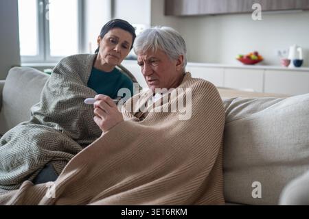 Erschöpfte ältere Menschen leiden an saisonaler Erkältungskrankheit, Ehemann kontrolliert die Temperatur Stockfoto