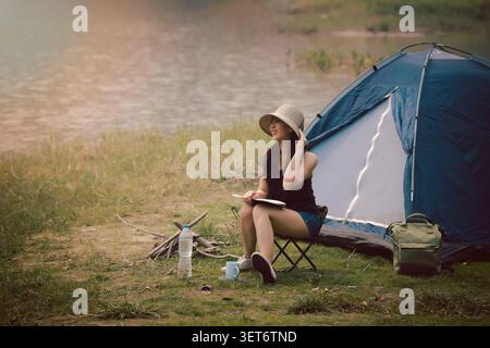 Glückliche Frau, die auf dem Campingstuhl sitzt und ein Buch vor dem Campingzelt auf der Wiese in der Nähe des Sees liest. Freizeitaktivitäten und Freizeitaktivitäten im Freien Stockfoto