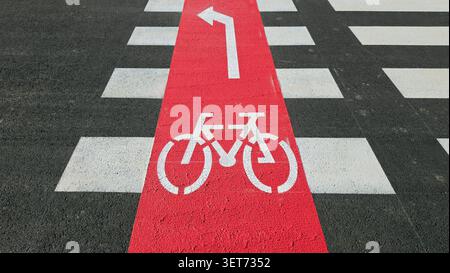 Ein hoher Blickwinkel zeigt einen rot lackierten Fahrradweg, der eine schwarze Asphaltstraße mit weißen Fußgängerzebrastreifen überquert und ein schabloniertes Fahrrad zeigt Stockfoto