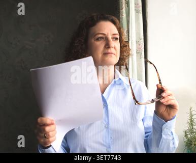 Reife Frau, die Dokumente und Brillen hält und in weichem natürlichem Licht nachdenklich durch ein Fenster schaut Stockfoto