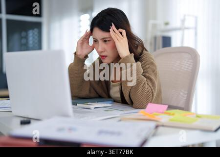 Junge asiatische Frau in beigefarbener Strickjacke, die Schläfen berührt und Stress und Kopfschmerzen am Arbeitsplatz mit Computer und zeigt Stockfoto