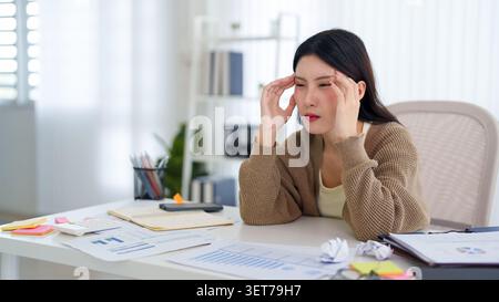 Gestresste junge asiatische Geschäftsfrau, die sich beim Berühren von Tempeln überwältigt fühlte, während sie am Schreibtisch an Berichten arbeitete Stockfoto