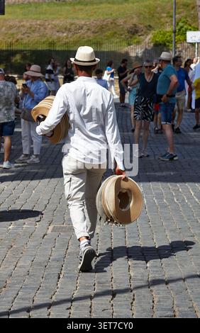Rom, Italien - 15. Juni 2026: Mann in weißem Hemd und beiger Hose hält mehrere Strohhüte, während er durch einen überfüllten Markt in Rom spaziert Stockfoto