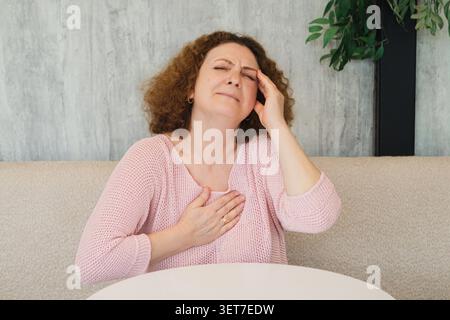 Frau mittleren Alters, die Brustschmerzen und Kopfschmerzen erlebt, während sie zu Hause sitzt und sich unwohl und Stress fühlt Stockfoto