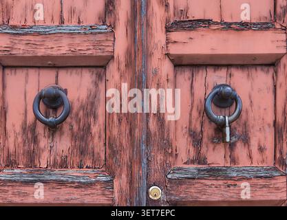 Nahaufnahme einer verwitterten Holztür mit abblätternder orangefarbener Farbe, mit antiken Metallklopfern und einem Vorhängeschloss auf der rechten Seite Stockfoto