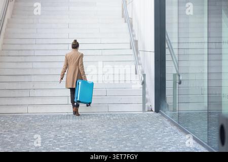 Eine Frau in einem braunen Mantel geht eine breite Steintreppe an einem modernen Bahnhof hinunter und zieht ihren blauen Koffer zu ihrem Abfahrtsteig. Stockfoto