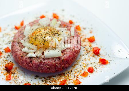 Rohe Rinderpastete mit rohem Eigelb drauf. Gehackte Zwiebeln und rote Paprikaflocken umgeben ihn. Schwarzer Pfeffer und Gewürze garnieren das Gericht. Serviert auf Clea Stockfoto