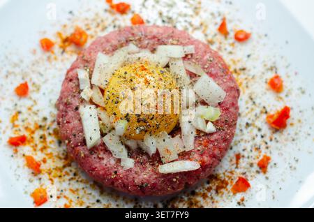 Rohe Rinderpastete mit rohem Eigelb drauf. Gehackte Zwiebeln und rote Paprikaflocken umgeben ihn. Schwarzer Pfeffer und Gewürze garnieren das Gericht. Serviert auf Clea Stockfoto