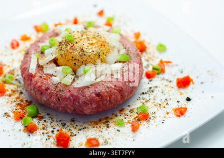 Rohe Rinderpastete mit rohem Eigelb drauf. Gehackte Zwiebeln und rote Paprikaflocken umgeben ihn. Schwarzer Pfeffer und Gewürze garnieren das Gericht. Serviert auf Clea Stockfoto