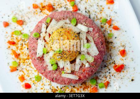 Rohe Rinderpastete mit rohem Eigelb drauf. Gehackte Zwiebeln und rote Paprikaflocken umgeben ihn. Schwarzer Pfeffer und Gewürze garnieren das Gericht. Serviert auf Clea Stockfoto