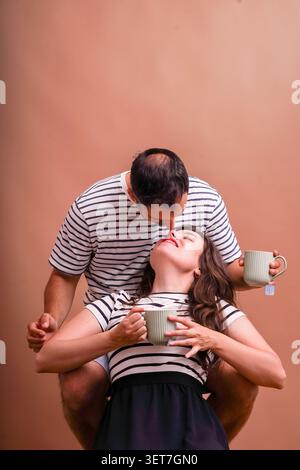 Ein junges Paar teilt sich einen liebevollen Kuss, während er heiße Getränke in gestreiften Hemden trinkt Stockfoto