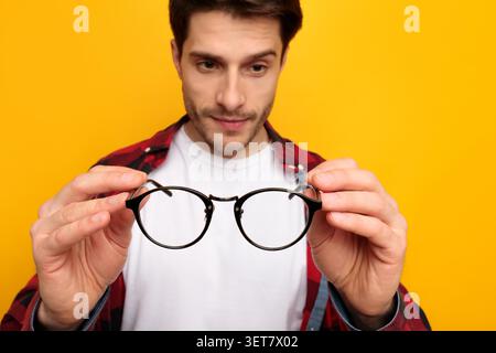 Mann Untersucht Schwarze Runde Brille In Händen Mit Fokussiertem Ausdruck, Gelber Hintergrund Stockfoto