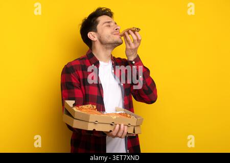 Glücklicher Junger Kaukasischer Mann, Der Pizzaschnitzel Isst, Pizzaschachtel Hält, Gelber Hintergrund Stockfoto