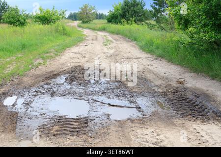 Eine große Schlammpfütze mit Traktorreifen liegt auf einer ländlichen Feldstraße durch Wald und Wiesen zu einem Bauernhof nach Regen. Windung nasser Tonweg mit Wasser Stockfoto
