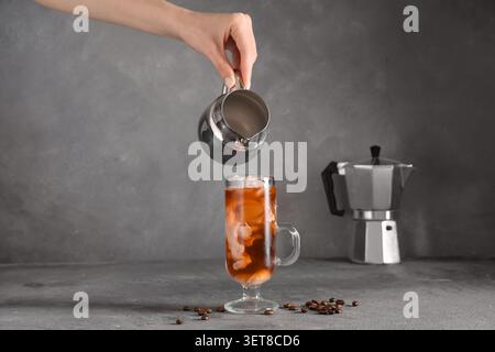 Creme von Hand in Eiskaffee mit Eiswürfeln in einem Glasbecher, Kaffeebohnen und Mokakanne auf grauem Hintergrund gießen Stockfoto
