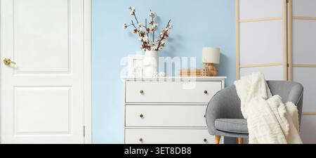Moderne Inneneinrichtung mit weißer Kommode, Baumwollzweigen in Vase und gemütlichem grauen Sessel vor blauer Wand Stockfoto