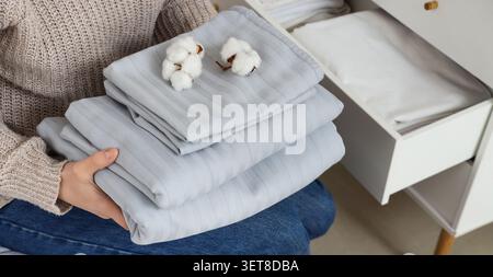 Frau, die einen Stapel frisch gewaschener Baumwollbettwäsche mit dekorativen Baumwollbollen hält, Wohnorganisation und Komfort Stockfoto