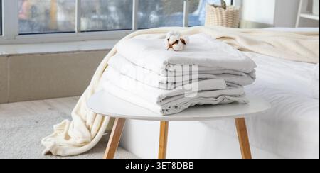 Stapel frisch gewaschene weiße Baumwollbettwäsche mit Baumwoll-Boll auf einem kleinen Beistelltisch in einem hellen Schlafzimmer, eine Baumwoll-Boll aus natürlicher Baumwolle liegt darauf Stockfoto