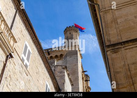 Blick auf einen mittelalterlichen Burgturm aus Stein, der eine rote und blaue Flagge zwischen alten Gebäuden vor einem blauen Himmel in Uzes, Frankreich, weht Stockfoto