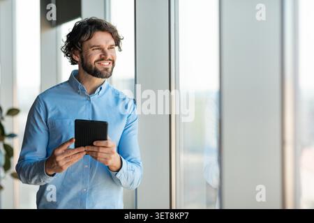 Glücklicher Kaukasischer Männlicher Profi, Der Das Tablet Beim Anlehnen Gegen Das Fenster Im Modernen Büro Verwendet Stockfoto