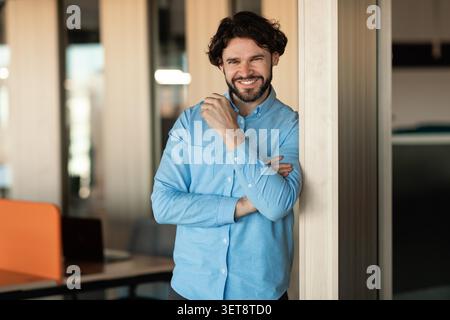 Lächelnder Kaukasischer Mann, Der Sich An Die Wand Lehnt In Moderner Büroumgebung, Geschäftskonzept Stockfoto