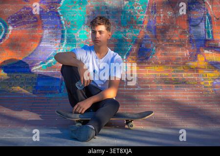 Ein junger Mann, der auf seinem Skateboard sitzt und mit dem Rücken an einer bunten Wand ruht. Stockfoto