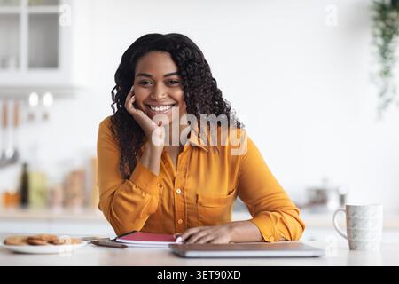 Lächelnde Junge Afroamerikanische Frau Sitzt Am Küchentisch Mit Laptop Und Kaffee Stockfoto
