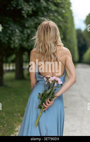 Eine Frau läuft einen von Bäumen gesäumten Weg entlang. Sie hält einen Haufen Blumen hinter sich. Die Sonne scheint auf die grünen Blätter und den Boden. Die Szene-Show Stockfoto