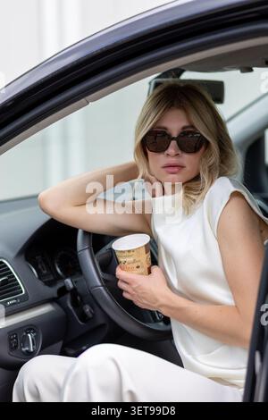 Eine Frau sitzt bequem in einem Auto mit einem Arm auf dem Lenkrad. Sie hält eine Kaffeetasse in der Hand und trägt eine Sonnenbrille. Ihr leichtes Tuch Stockfoto