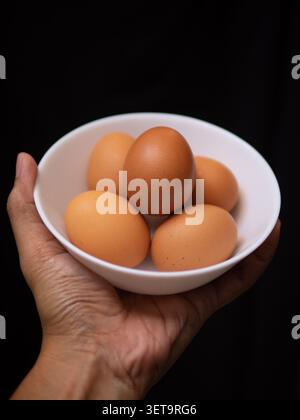 Fünf Hühnereier auf einer weißen Schüssel, gehalten von einer linken Hand auf schwarzem Hintergrund Stockfoto
