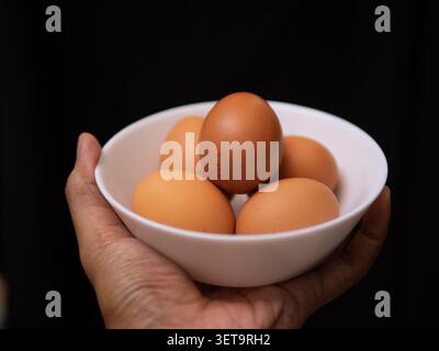 Fünf Hühnereier auf einer weißen Schüssel, gehalten von einer linken Hand auf schwarzem Hintergrund Stockfoto