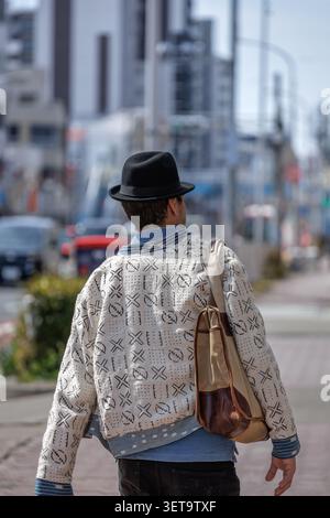 Rückansicht eines modischen Mannes in einer cremefarbenen Jacke und einem schwarzen Fedorahut, der auf einem Bürgersteig der Stadt mit einer Canvas-Umhängetasche läuft. Stockfoto