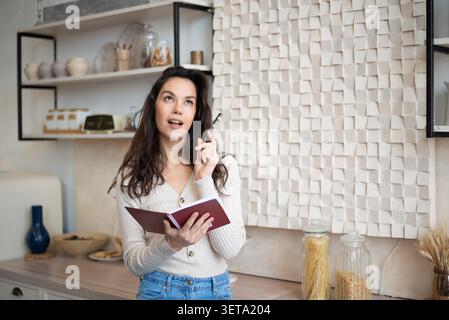 Nachdenkliche Junge Frau Schreibt In Notizbuch Zu Hause, In Der Küche. Lifestyle-Konzept Stockfoto