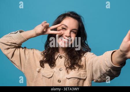 Happy Young Woman Lächelt Und Posiert Mit Peace Sign In Studio, Blauer Hintergrund Stockfoto