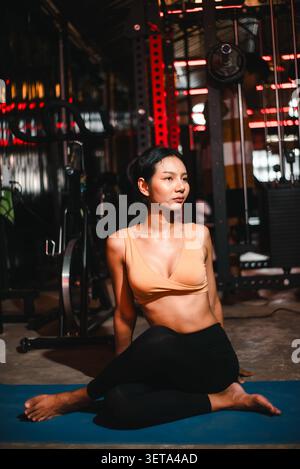 Athletische asiatische Frau, die in einem dunklen Fitnessstudio auf einer blauen Yogamatte sitzt und nach dem Training eine sitzende Drehung übt, Flexibilität, Mobilität, Erholung zeigt, achtsam Stockfoto