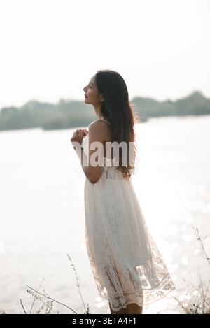 Junge asiatische Frau in einem weißen Spitzenkleid, die an einem hellen See mit gefesselten Händen steht, Dankbarkeit, Hoffnung, friedliche Natur, sanftes Sonnenlicht zeigt, achtsam Stockfoto