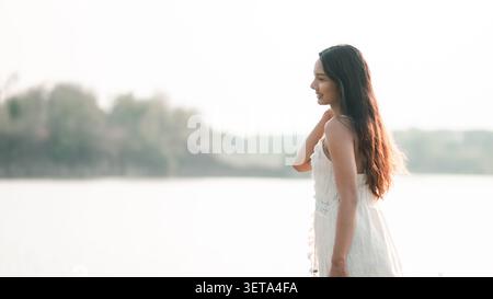 Junge asiatische Frau in einem weißen Spitzenkleid, lächelnd neben einem ruhigen See bei weichem Tageslicht, mit friedlichem Reisen, natürlicher Schönheit, Freiheit, Outdoor-Lifestyle Stockfoto