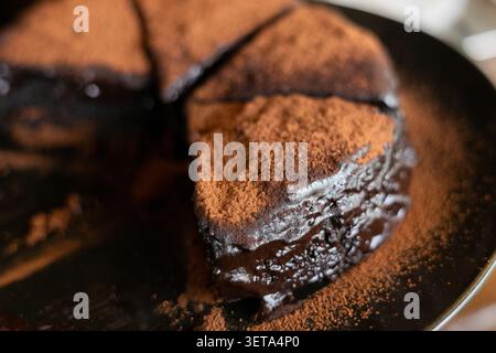 Reichhaltiger, feuchter Schokoladenkuchen mit glänzender Schokoladenganache und Kakaopulver, in Scheiben geschnitten und auf dunklem Teller serviert, um ein dekadentes und verwöhnendes DE zu kreieren Stockfoto