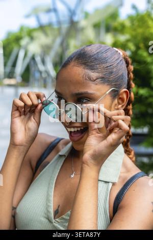 Birassische junge Frau, die eine runde Sonnenbrille senkt, verspielt mit einem breiten Lächeln blickt und Glück und Freude ausdrückt, während sie einen lebhaften Sommertag genießt Stockfoto