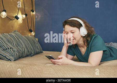 Frau, die auf dem Bett liegt, Kopfhörer trägt, lächelt und ein Handy benutzt Stockfoto