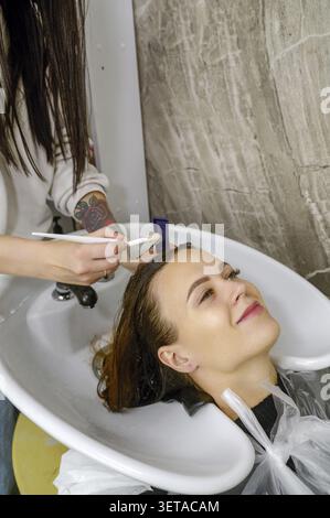 Eine Nahaufnahme der Hand eines Friseurs und eines zufriedenen Kunden, der ein Peeling, eine Maske oder eine Farbe auf das Haar in einem Salon aufträgt. Friseur- und Haarpflegekonzept. Hai Stockfoto