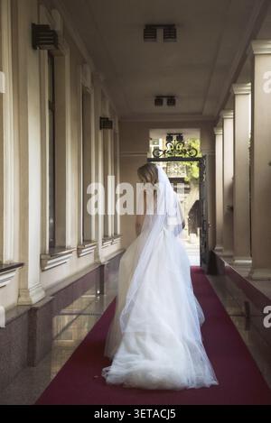 Wunderschöne blonde Braut in einem wunderschönen Hotel, in einem Bogen in einem langen Hochzeitskleid. Braut, Blick von hinten, sie hat gewelltes blondes Haar, offene Schultern. Stockfoto