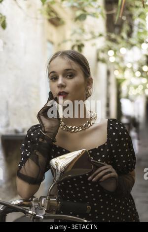 Porträt einer niedlichen und stilvollen jungen Frau in einem schwarzen Kleid mit weißen Polka Dots und schwarzen Handschuhen neben einem klassischen Roller in einer Altstadt. Vintage s Stockfoto