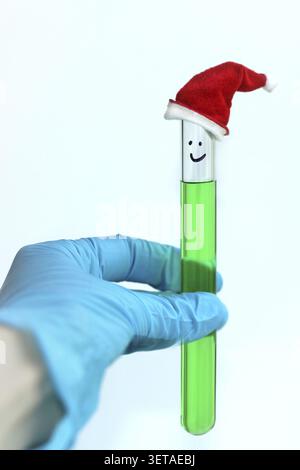 Eine Hand mit blauen Handschuhen hält ein Reagenzglas mit einer grünen Lösung und einer dekorativen roten Kappe im Chemielabor. Neujahrsstimmung bei der Arbeit Stockfoto