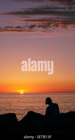 Mann Silhouette sitzt auf Felsen und beobachtet den Sonnenuntergang über dem Meer vertikal. Stockfoto