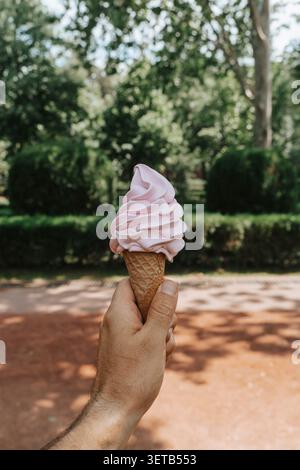 Hand hält einen Schuss rosafarbenes Eis in einer Waffelschale vor einem sanft verschwommenen Hintergrund eines sonnendurchfluteten Parks und fängt die erfrischende Essenz eines Sommertages ein. Stockfoto