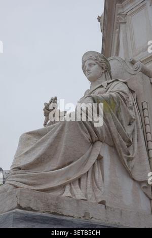 Klassische Steinstatue in fließenden Gewändern mit Kreuz an der Fassade des historischen Gebäudes, flacher Blick vor grauem Himmel Stockfoto