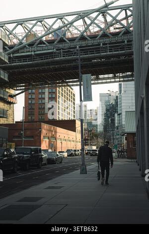 New York, New York, Vereinigte Staaten - 14. März 2025: Blick auf die Manhattan Bridge, die über einer Straße thront und von einem Fußgänger auf dem Bürgersteig, Park, spaziert Stockfoto