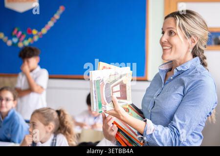 Freundliche Lehrerin, die einen Stapel Bücher hält und sich während eines Unterrichts in einem Grundschulklassen mit Schülern beschäftigt. Bildung, Lernen, te Stockfoto