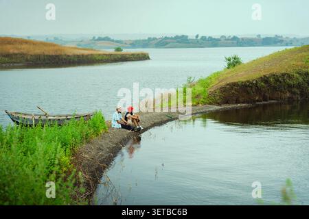 Zwei Personen sitzen an einem ruhigen See mit einem kleinen Boot in der Nähe, umgeben von grünem Gras und sanften Hügeln. Stockfoto
